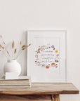 A Weekend Full Of Little Things fine art print shown in a slim white frame with a white mount, leaning on a rustic wooden table beside a round white vase of dried bunny tail grasses. Frame not included.