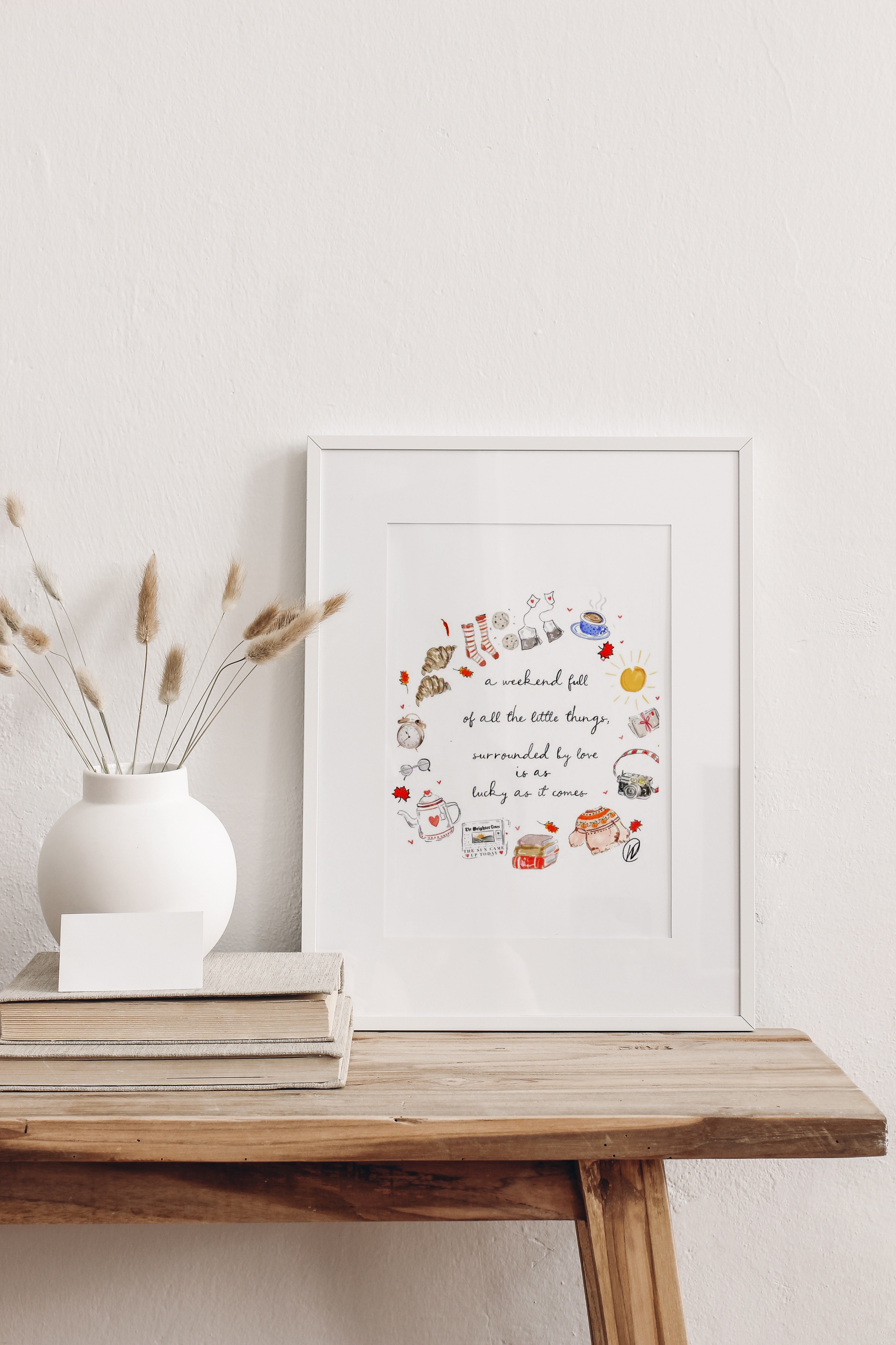 A Weekend Full Of Little Things fine art print shown in a slim white frame with a white mount, leaning on a rustic wooden table beside a round white vase of dried bunny tail grasses. Frame not included.