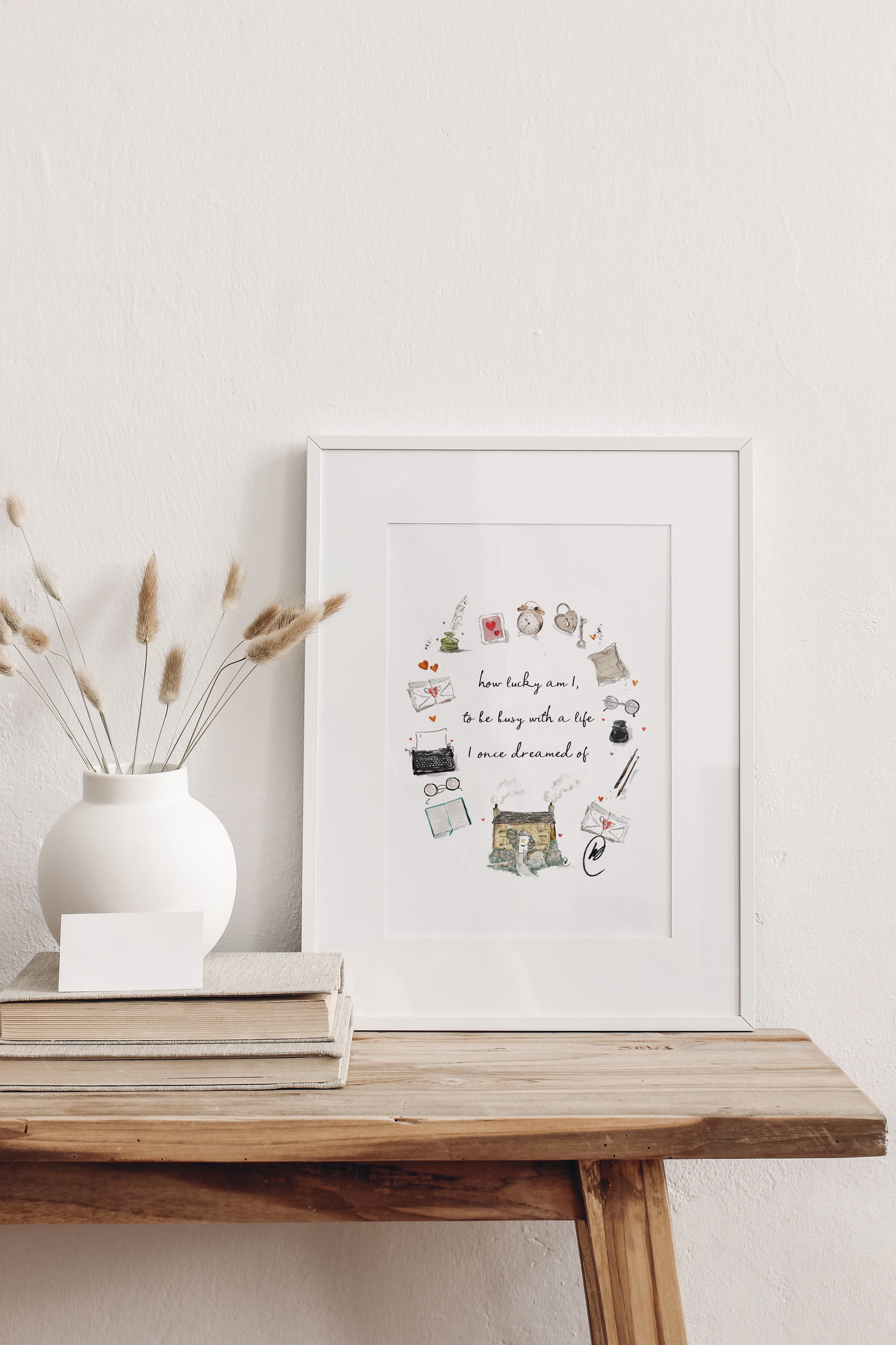 The 'how lucky am I, to be busy with a life I once dreamed of' cottage wreath illustration shown as a framed print in a slim white frame with a white mount, leaning on a rustic wooden table beside a white vase of dried grasses. Frame not included.