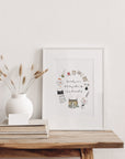 The 'how lucky am I, to be busy with a life I once dreamed of' cottage wreath illustration shown as a framed print in a slim white frame with a white mount, leaning on a rustic wooden table beside a white vase of dried grasses. Frame not included.
