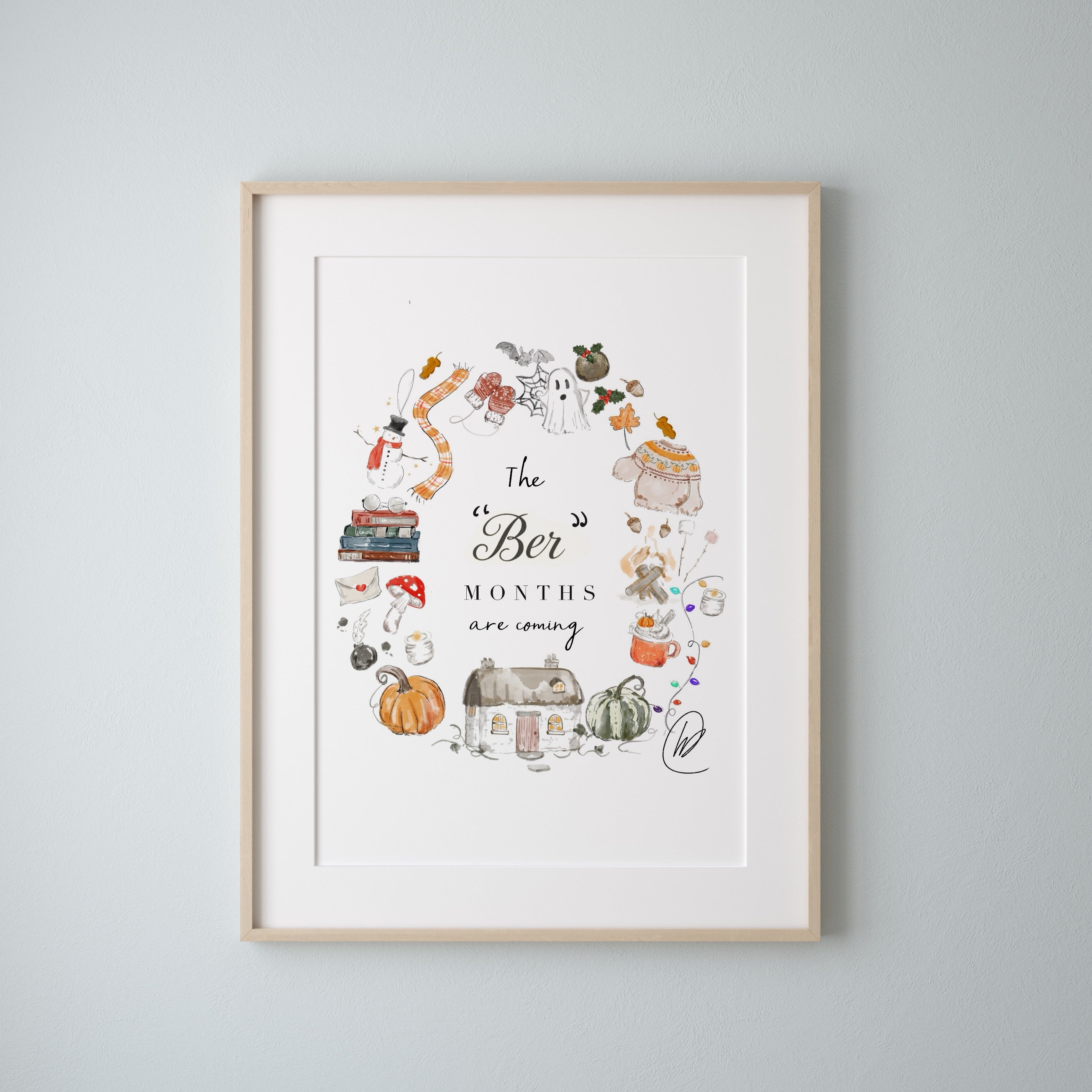 The Ber Months Are Coming print shown in a light oak frame with a white mount on a pale grey blue wall, the watercolour wreath of autumn and winter things sitting inside the mount window. Frame not included.