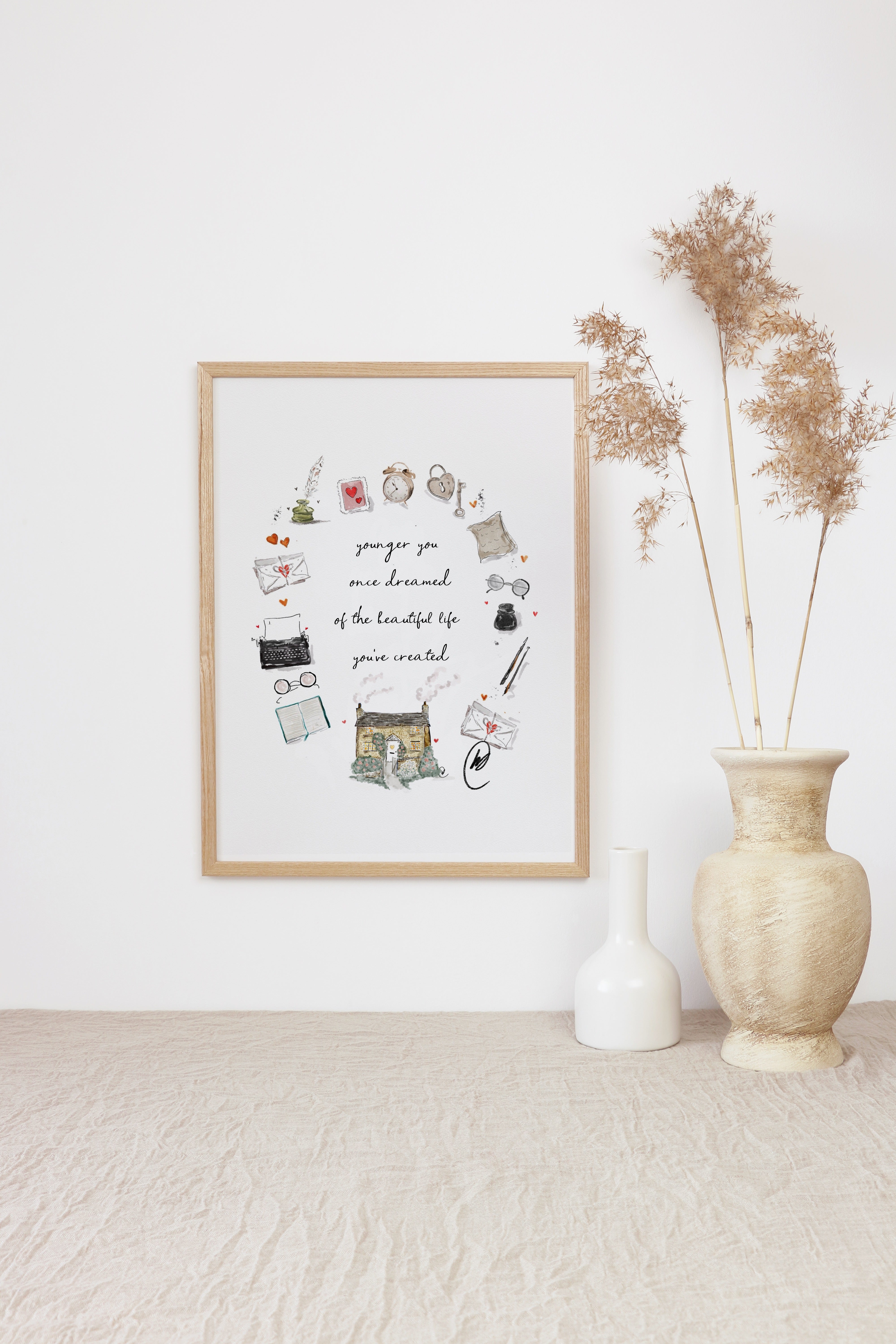 The 'younger you once dreamed of the beautiful life you've created' cottage wreath illustration shown as a framed print in a light oak frame, leaning against a white wall beside tall dried pampas grasses in a pale vase. Frame not included.